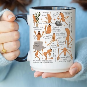 Puede incluir: Taza de cerámica blanca con asa y borde negros, con ilustraciones de dibujos animados de personajes de pasta con diversas expresiones y frases. La taza incluye texto como "RIGATONI" y "Estoy ligeramente irritado."