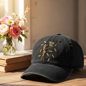 May include: Black baseball cap with a curved brim. The cap features a gold graphic of a person holding pasta and the words "RIGATONI PASTA". The design is surrounded by gold pasta shapes. The cap is sitting on a wooden surface.