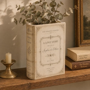 May include: A cream-colored ceramic book-shaped vase featuring decorative borders and text that reads: Our Greatest Chapter, A Love Story, Written by Sophia & Ethan, Est. 04.12.2025, Every Page Leads Us Home. The spine displays Sophia & Ethan.