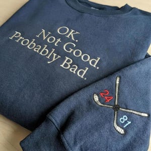 Puede incluir: Sudadera azul marino con texto bordado en blanco que dice "OK. Not Good. Probably Bad." La manga presenta palos de hockey cruzados con los números 24 y 81 bordados en rojo y azul.