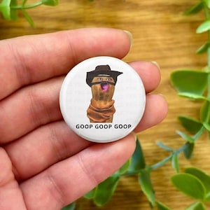 Può includere: Un bottone bianco con un cartone animato di una nocciolina con cappello da cowboy e sciarpa. La nocciolina ha un'espressione sorpresa con la bocca aperta. Il testo "GOOP GOOP GOOP" è stampato in nero sotto.