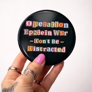 Puede incluir: Un botón redondo negro con el texto "Operation Epstein War - Don't Be - Distracted" en letras coloridas recortadas. El botón está sostenido por una mano con esmalte de uñas rosa y varios anillos.