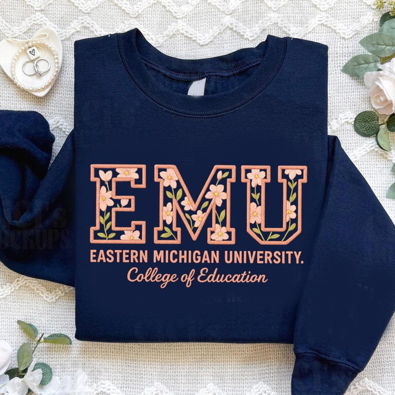 May include: A navy blue sweatshirt featuring the letters "EMU" filled with a floral pattern. The text "EASTERN MICHIGAN UNIVERSITY. College of Education" is below in peach. The garment rests on a white, textured background.