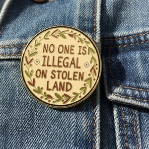 May include: A round enamel pin with the text "NO ONE IS ILLEGAL ON STOLEN LAND" in brown lettering. The pin has a gold border and is decorated with green and brown leaves. The pin is attached to a blue denim jacket.