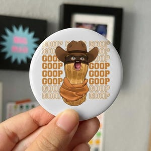 Può includere: Un bottone rotondo bianco con un cartone animato di una nocciolina con un cappello da cowboy, una maschera per gli occhi e una sciarpa. La nocciolina ha la bocca aperta e un'espressione sorpresa. La parola "GOOP" è ripetuta.