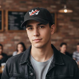 Pu&ograve; includere: Cappellino da baseball nero con logo rosso e bianco. Il cappellino &egrave; indossato da una persona che indossa una maglietta grigia e una giacca di jeans nera. Lo sfondo comprende un muro di mattoni e persone sedute ai tavoli.