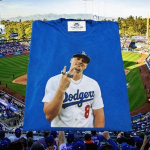 May include: A blue t-shirt with a white label that reads "THE HEARTLESS HOME". The shirt displays a graphic of a baseball player in a Dodgers uniform. The player is wearing a Dodgers cap and multiple rings.