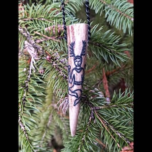 May include: A pendant necklace featuring a carved antler pendant with a black ink drawing of a figure with antlers. The pendant hangs from a black chain and is set against a backdrop of green pine needles.