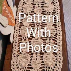 May include: A crocheted table runner with a beige and pink color scheme. The runner features a repeating pattern of hearts and has a scalloped edge. The text "Pattern With Photos" is superimposed on the image.