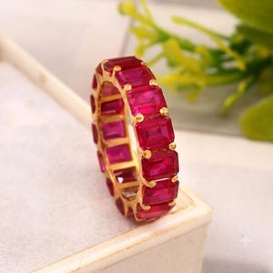 May include: An eternity band ring featuring a continuous row of vibrant, rectangular-cut, deep pinkish-red gemstones set in a polished yellow gold metal frame. The stones are arranged in a repeating pattern around the entire circumference of the ring.