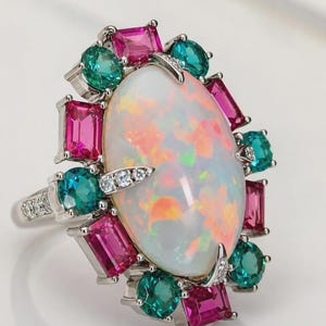 May include: A silver-toned ring featuring a large oval opal center stone with a play-of-color. The opal is surrounded by a halo of alternating emerald-cut pink stones and round-cut teal stones. The band and prongs are accented with small white stones.
