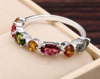 May include: A silver ring featuring a row of multi-colored faceted gemstones, including oval-cut red, yellow, and green stones set in a polished metal band. The stones are arranged in a sequence along the top half of the circular ring structure.