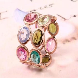 May include: A rose gold ring featuring a band of multiple oval-cut gemstones in various colors, including pink, light blue, olive green, and teal. The stones are set in individual metal bezels arranged in a circular, multi-row pattern.