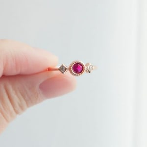 May include: A delicate rose gold ring featuring a central round, vibrant red gemstone, flanked by two smaller, diamond-shaped accents. The ring's band is slender, and the design is simple yet elegant, suitable for everyday wear.