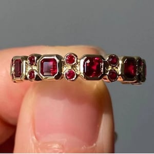 Könnte beinhalten: Ein Goldring mit einer Reihe roter Rubin-Edelsteine. Der Ring hat eine Mischung aus quadratischen und runden Steinen.