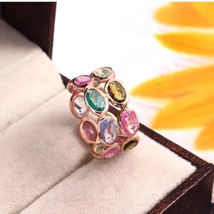 May include: A rose gold band ring featuring two rows of oval-cut multi-colored gemstones. The stones include shades of pink, green, blue, and yellow, all set in a metallic frame that encircles the finger with a continuous pattern of faceted gems.