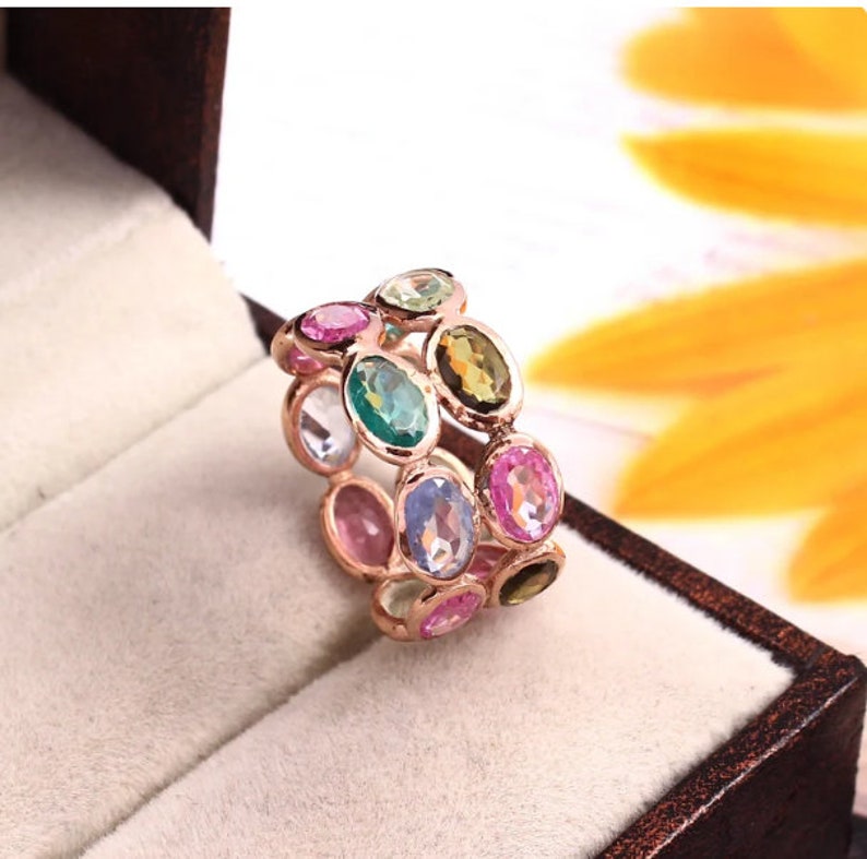 May include: A rose gold band ring featuring two rows of oval-cut multi-colored gemstones. The stones include shades of pink, green, blue, and yellow, all set in a metallic frame that encircles the finger with a continuous pattern of faceted gems.