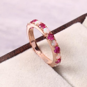 May include: A rose gold ring featuring a row of alternating red gemstones and white opals. The ring is set against a soft, neutral background, highlighting the vibrant colors of the stones.