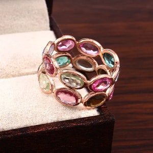 May include: A multi-stone ring featuring a rose gold band set with numerous oval-cut gemstones in various colors, including shades of pink, green, and blue. The stones are arranged in a continuous, linked pattern around the entire circumference of the ring.
