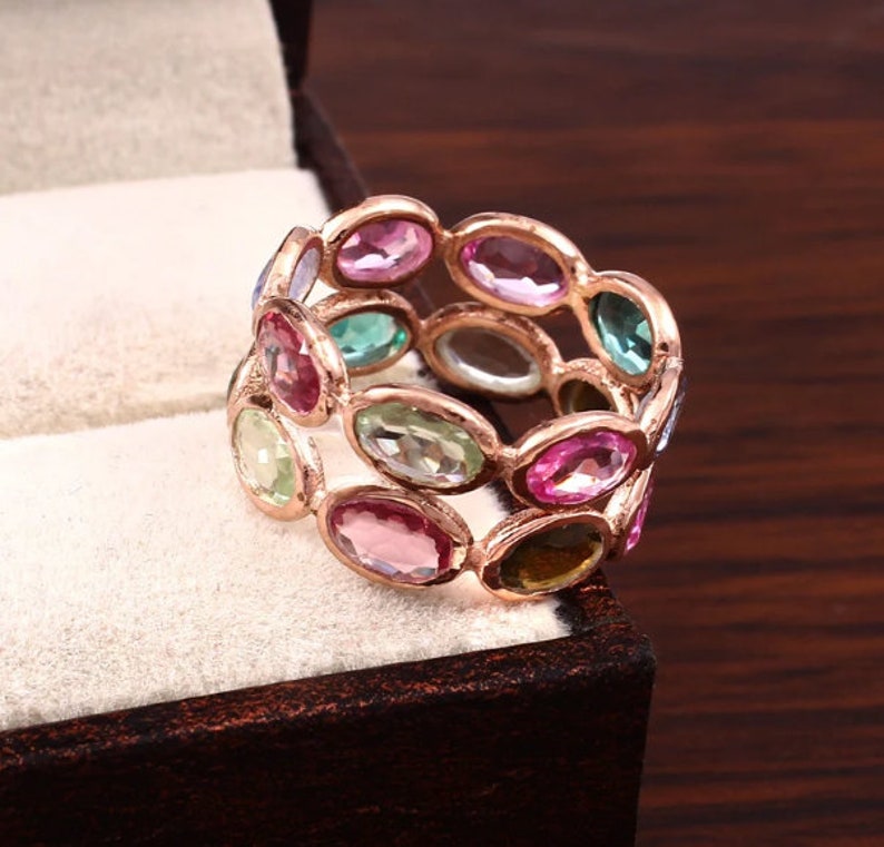 May include: A multi-stone ring featuring a rose gold band set with numerous oval-cut gemstones in various colors, including shades of pink, green, and blue. The stones are arranged in a continuous, linked pattern around the entire circumference of the ring.