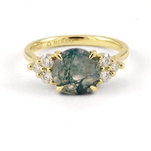 May include: A gold ring with a green agate gemstone and small diamonds set around it. The ring is inscribed with the word "MERAKI".