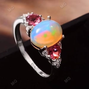 May include: A silver ring featuring a large oval opal gemstone, surrounded by smaller pink and white gemstones.