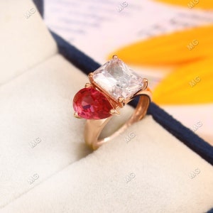 Può includere: Un anello d'oro con una grande pietra preziosa quadrata trasparente e una piccola pietra preziosa rossa a forma di lacrima.