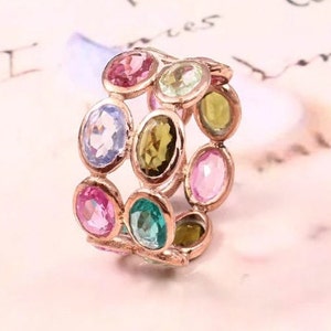 May include: A rose gold band ring featuring multiple oval-cut gemstones in various colors, including pink, light blue, green, and yellow. The stones are set in individual metal bezels arranged in a multi-row pattern around the circumference.