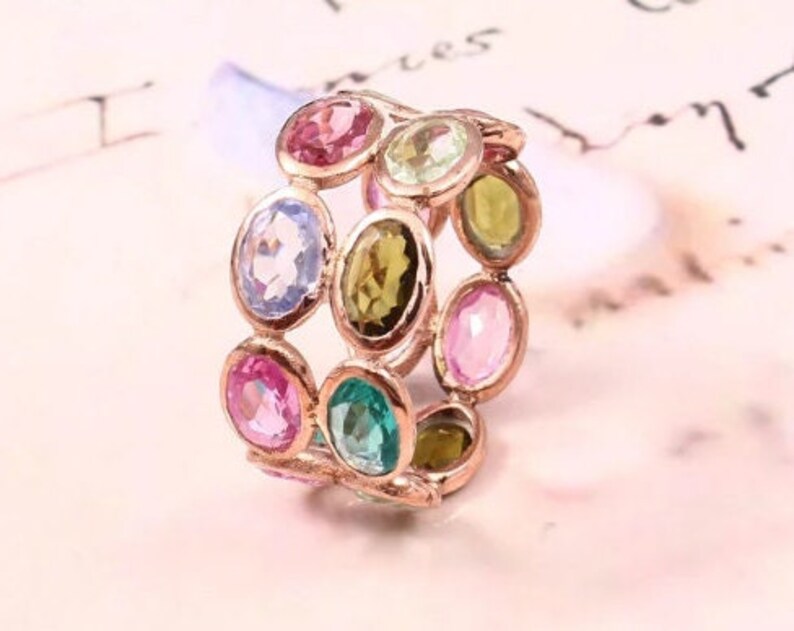 May include: A rose gold band ring featuring multiple oval-cut gemstones in various colors, including pink, light blue, green, and yellow. The stones are set in individual metal bezels arranged in a multi-row pattern around the circumference.
