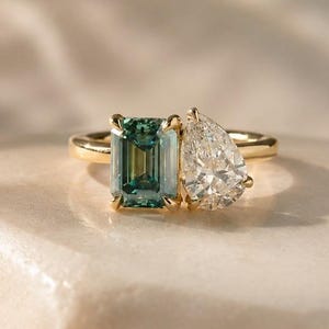 Puede incluir: Un anillo de oro con dos piedras preciosas: una piedra rectangular de color verde azulado y una piedra clara en forma de pera. El anillo tiene una banda delgada, sobre un fondo neutro. Las piedras están engastadas en oro.