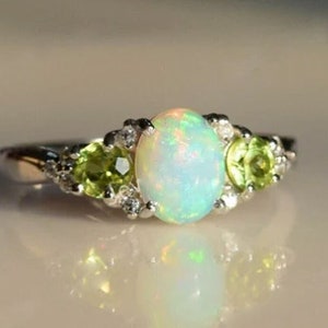 Puede incluir: Un anillo de plata con una gran piedra de ópalo ovalada rodeada de piedras preciosas más pequeñas de color verde y transparente.