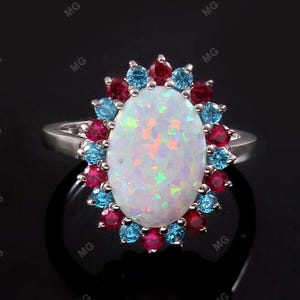 May include: A silver ring featuring an oval, iridescent opal center stone, surrounded by a halo of alternating red and blue gemstones. The ring is set against a black background, highlighting the vibrant colors of the gemstones.