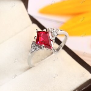 May include: A silver-toned ring featuring a central kite-shaped red gemstone held by four prongs. The band is accented on both sides by clusters of three small, clear, round-cut stones set in a triangular arrangement, creating a symmetrical design.