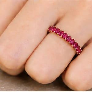 May include: A gold eternity band ring featuring a continuous row of small, round, vibrant red gemstones set in a shared prong setting. The ring encircles the finger, showcasing a uniform arrangement of stones that create a bright, saturated color.
