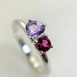 May include: A silver ring featuring a purple amethyst and a red garnet gemstone.