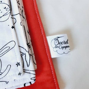 Puede incluir: Se ve una pila de art&iacute;culos de tela blanca con dibujos lineales negros de personajes con tem&aacute;tica espacial, incluyendo un conejo y estrellas. Los art&iacute;culos est&aacute;n sobre un fondo rojo. Tambi&eacute;n se ve una etiqueta blanca con el texto "Secret des Cigognes".