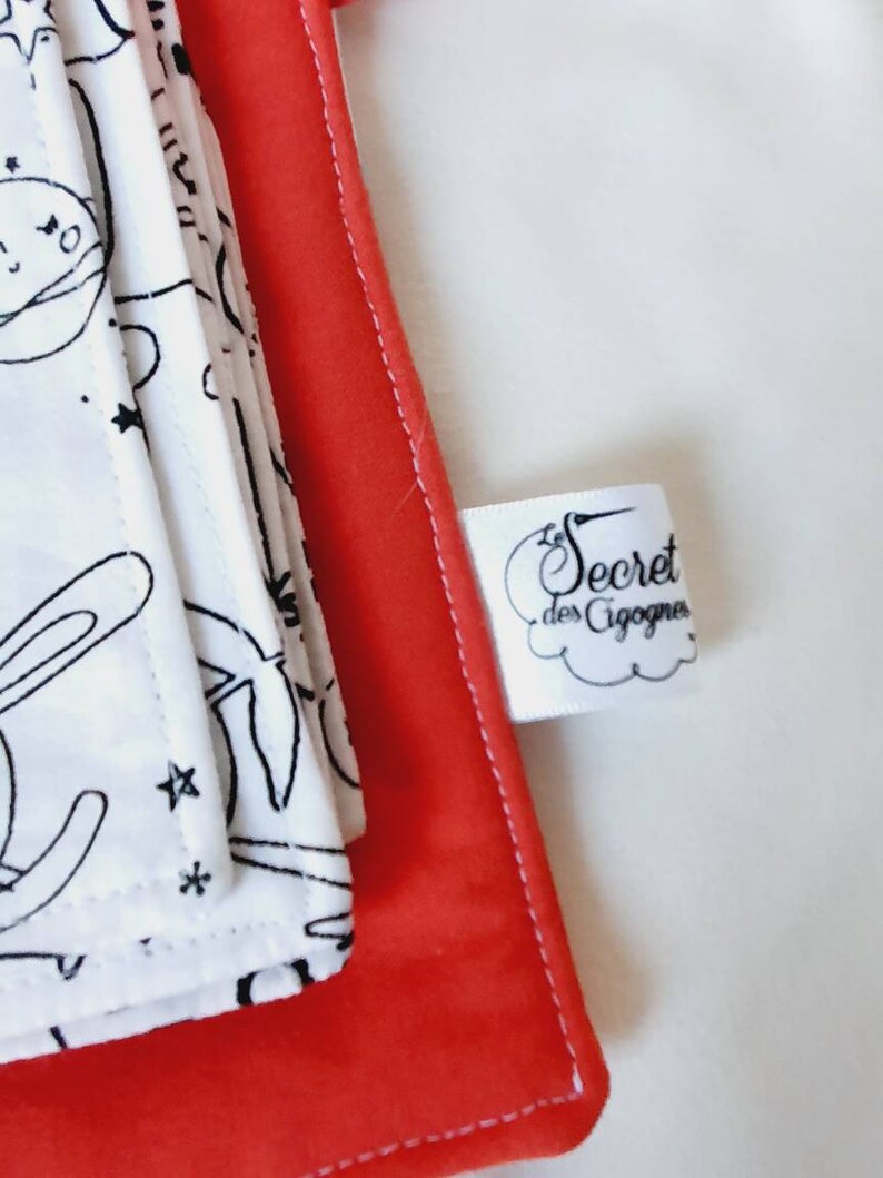 Puede incluir: Se ve una pila de art&iacute;culos de tela blanca con dibujos lineales negros de personajes con tem&aacute;tica espacial, incluyendo un conejo y estrellas. Los art&iacute;culos est&aacute;n sobre un fondo rojo. Tambi&eacute;n se ve una etiqueta blanca con el texto "Secret des Cigognes".