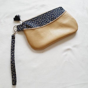 Puede incluir: Un pequeño clutch de cuero marrón con un forro de tela estampado floral azul y blanco y una correa de muñeca de tela a juego.