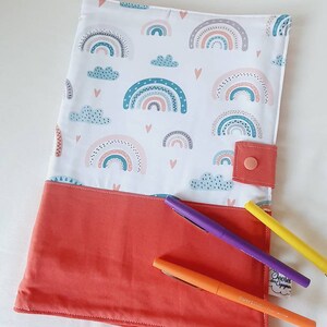 Puede incluir: Una funda de tela blanca con una secci&oacute;n inferior de color coral. La parte superior presenta un patr&oacute;n de arco&iacute;ris, nubes y corazones de colores. Se ve un cierre a presi&oacute;n de color coral. Tres bol&iacute;grafos en morado, naranja y amarillo est&aacute;n sobre la funda.