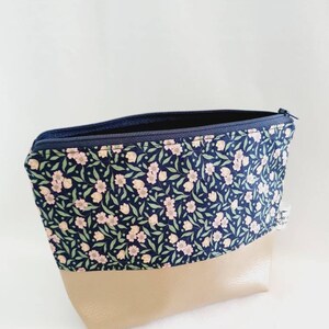 Puede incluir: Un neceser con estampado floral, cremallera azul marino y base beige. La parte superior de la bolsa es azul marino con un estampado de pequeñas flores rosas y hojas verdes. La parte inferior es de color beige liso.