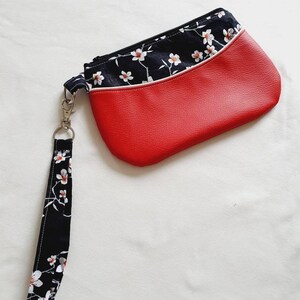 Puede incluir: Un bolso de mano rojo y negro con un estampado floral. El bolso de mano tiene un cuerpo rojo similar al cuero con una sección superior negra con flores blancas. Una correa de muñeca a juego está unida con un cierre plateado. El bolso de mano mide aproximadamente 23 cm de largo.