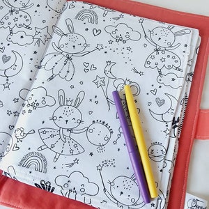 Puede incluir: Una cubierta de libro para colorear de color coral con un interior blanco con dibujos lineales negros de conejos, estrellas, lunas y nubes. Dos rotuladores, morado y amarillo, descansan sobre las p&aacute;ginas abiertas. El texto "Secret des Cigognes" es visible.