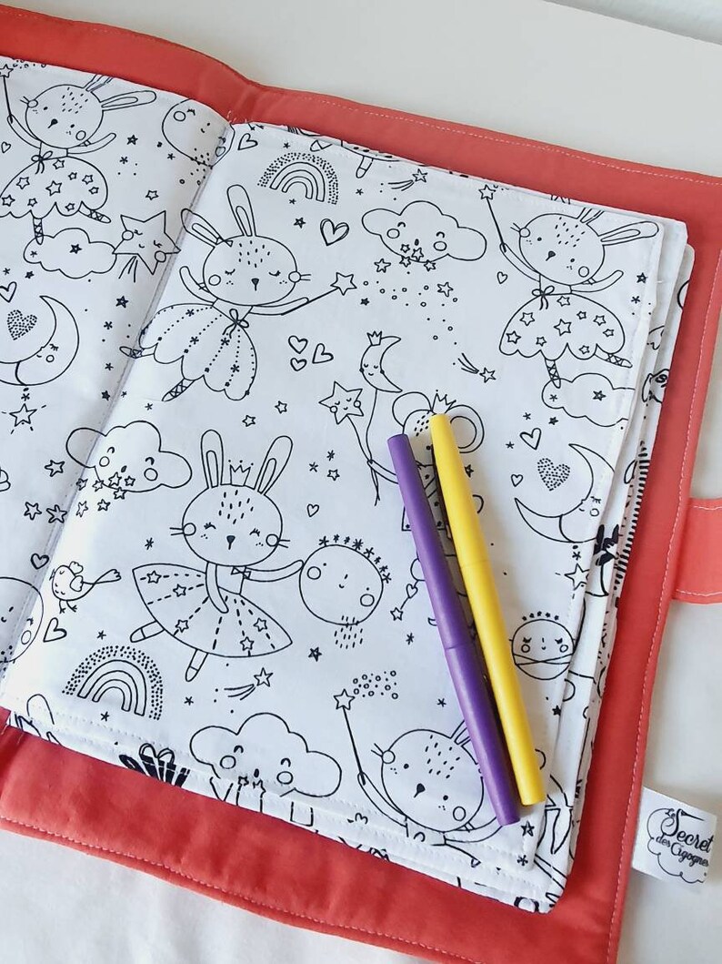 Puede incluir: Una cubierta de libro para colorear de color coral con un interior blanco con dibujos lineales negros de conejos, estrellas, lunas y nubes. Dos rotuladores, morado y amarillo, descansan sobre las p&aacute;ginas abiertas. El texto "Secret des Cigognes" es visible.