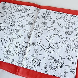 Puede incluir: Libro para colorear abierto con ilustraciones en blanco y negro de nutrias, peces, lib&eacute;lulas y plantas acu&aacute;ticas. El libro est&aacute; abierto sobre una superficie roja, con las p&aacute;ginas mostrando los dibujos detallados. El tema general es la naturaleza y la fauna.