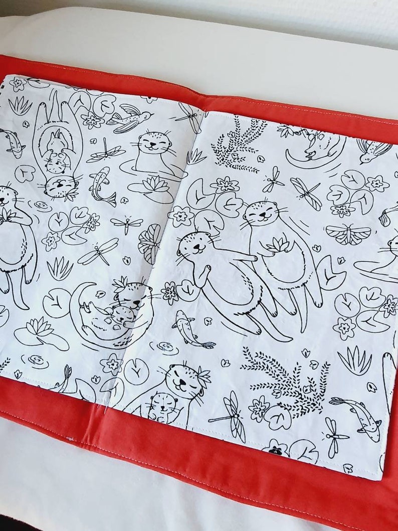 Puede incluir: Libro para colorear abierto con ilustraciones en blanco y negro de nutrias, peces, lib&eacute;lulas y plantas acu&aacute;ticas. El libro est&aacute; abierto sobre una superficie roja, con las p&aacute;ginas mostrando los dibujos detallados. El tema general es la naturaleza y la fauna.