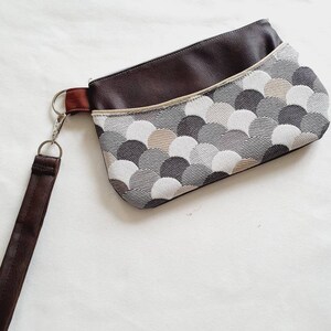 Puede incluir: Un pequeño bolso de mano de color marrón y gris con un patrón y una correa marrón. El bolso tiene una cremallera y una solapa.