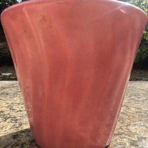 May include: A coral-colored ceramic planter with a glossy finish. The planter has a flared, conical shape, wider at the top and tapering towards the base. The surface shows subtle variations in color and texture.