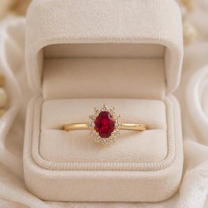 May include: A gold ring featuring an oval-cut red ruby centre stone surrounded by a halo of small, clear, round-cut diamonds. The ring is displayed inside a cream-coloured velvet jewellery box with a soft, padded interior lining.