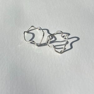 May include: Two silver rings with a flowing, irregular design. The rings are positioned on a white surface, creating shadows. The rings have a distinctive, organic shape, possibly handmade. The overall style is minimalist and contemporary. The rings are likely made of silver.