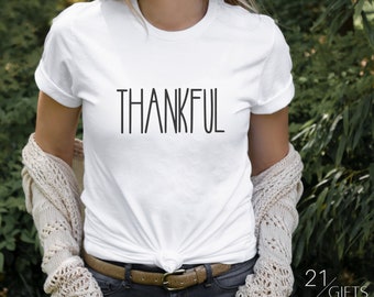 Puede incluir: Camiseta blanca con la palabra "THANKFUL" impresa en negro en la parte delantera. La camiseta la lleva puesta una persona con una chaqueta blanca de punto y un cinturón marrón.